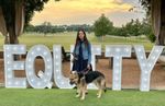 My Seeing Eye dog and I stand in front of a large sign. About four feet tall, the 3D letters spell EQUITY. Lightbulbs on each letter brighten the word. Behind them stand a row of trees, beyond which stretches a lush green field topped by clouds glowing in the setting sun.