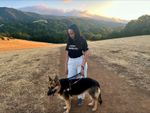I'm wearing a shirt that says &ldquo;Commit to Inclusion&rdquo; and smiling down at Mylo, a German Shepherd Seeing Eye dog. Behind us is a grass-covered hillside with forested hills in the distance.