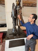 Mussie, a tall Eritrean American Deafblind man, stands beside the Helen Keller statue inside the U.S. Capitol. Young Helen has one hand on the handle of the water pump, and the other hand under the spout. Mussie studies Helen's hand beneath the water spout.