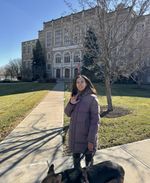 My guide dog Mylo and I stand on a path leading up to a stately stone building. I'm wearing a warm coat and signing ILY.