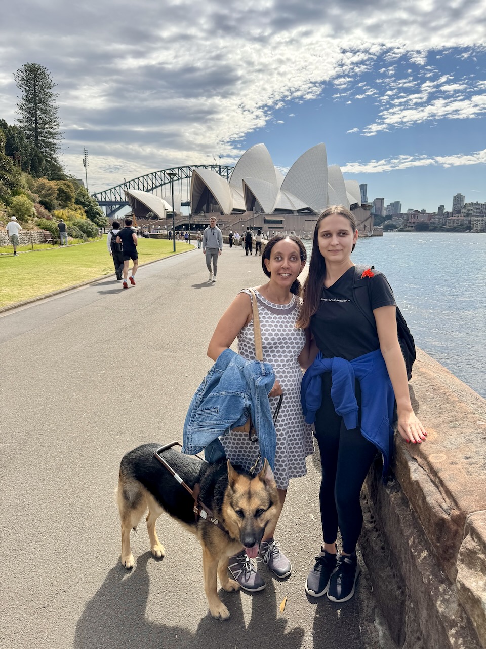 Video: Haben and Seeing Eye dog Mylo stand next to a young woman. They're all smiling, and behind them is the Sydney Opera House, the bright blue water of the harbor, and the bridge.