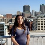 Haben stands on a rooftop on a sunny day with the Saint Louis skyline in the background. A large building with "U.S. Bank" on it is just over her shoulder. She is wearing a blue dress and smiling.