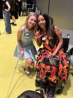 Two women smile as they lean toward each other for a picture. Keely Cat-Wells, a woman with light skin, long blonde hair,, wearing a sleeveless blue dress and white high heels, crouches beside Haben Girma. Haben, a woman with medium dark skin, long black hair, wearing a sleeveless multicolored dress and a support boot, is sitting in a wheelchair. She signs ILY with one hand while her other hand is on the Braille computer on her lap. Behind them is a large room with various people chatting.