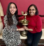 I'm sitting next to Laina, and between us is an oil lamp from Kerala — tall, brassy with a golden hue, and topped with a long-tailed bird.