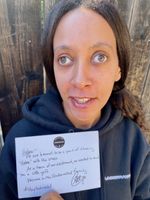 I'm wearing a black hoodie with the book club's name Underrated on the left side. I'm smiling and holding up the card. A dark brown fence is in the background.