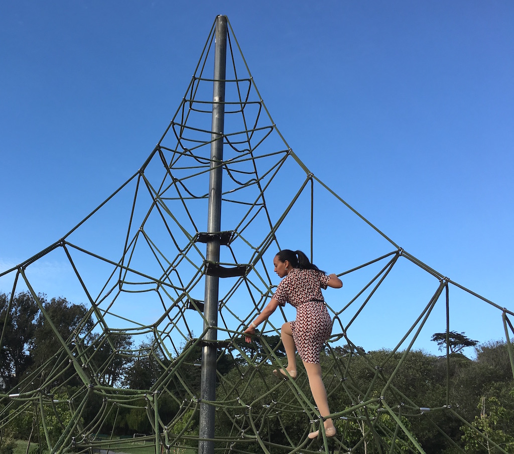 Haben climbs a large pyramid-shaped jungle gym that has multiple routes to the top. She is about two thirds of the way up the 20-foot high rope structure. Trees and blue skies are in the background.