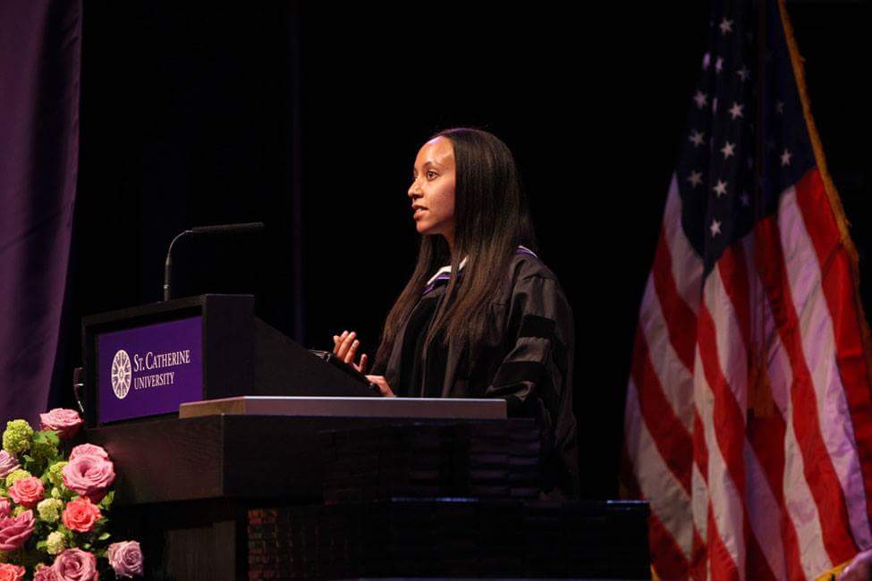 Haben is standing at the St. Catherine University podium wearing academic regalia, and behind her is a US flag.