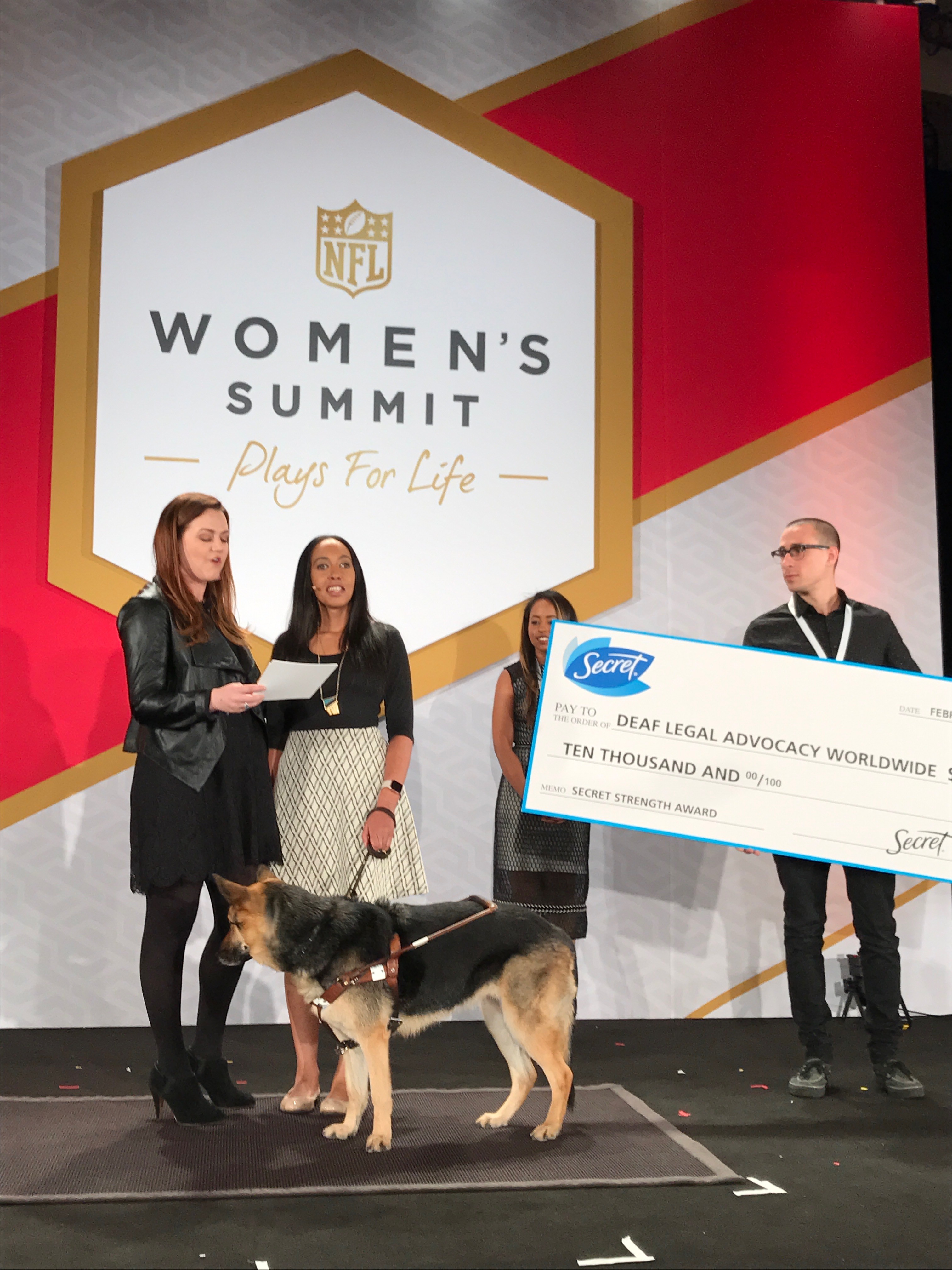 Secret's Nicole Summers reads from a page as Haben stands beside her. Maxine the Seeing Eye Dog is also on stage, head turned away from the audience. A man is holding a giant check with "Secret / Deaf Legal Advocacy Worldwide / $10,000 / Secret Strength Award.” Haben’s friend Mary is standing behind him. On stage is a red and gold banner, “NFL Women's Summit / Plays for Life." #NFLWomensSummit.
