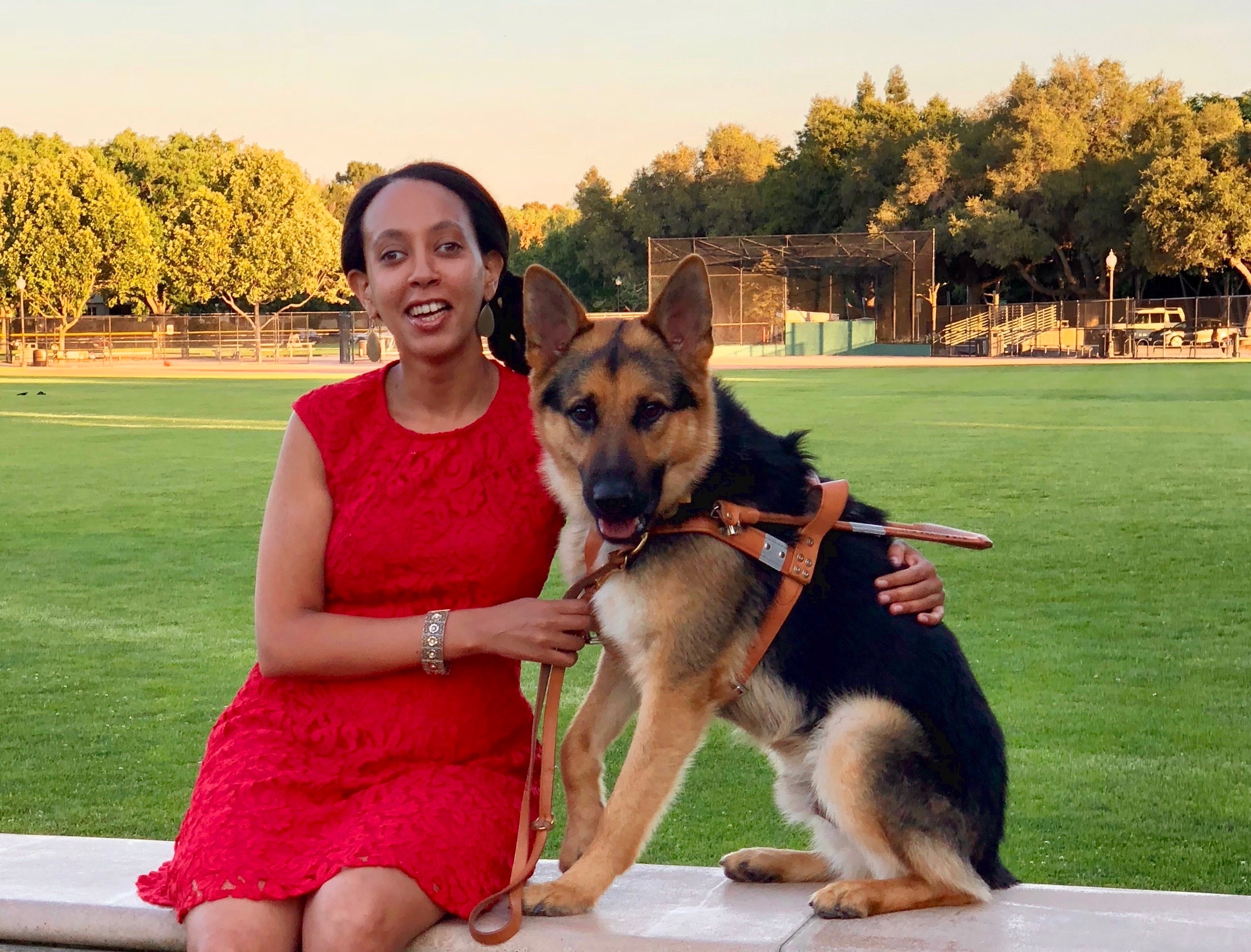 Haben and Mylo are sitting on a bench. Mylo is a black and tan medium-sized German Shepherd dog. He’s wearing a leather harness, and Haben is holding his leash. Behind them is a large grassy field with trees in the distance