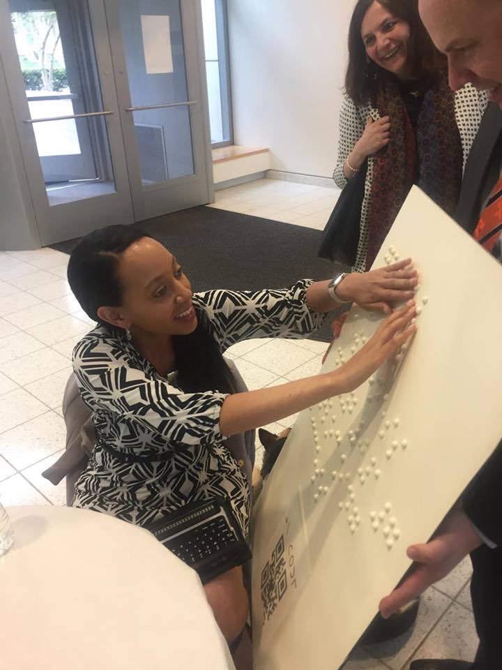 Damian Sian holds up a Princeton University jumbo braille poster as Haben reads it with two hands. She’s sitting on a chair with a braille computer on her lap