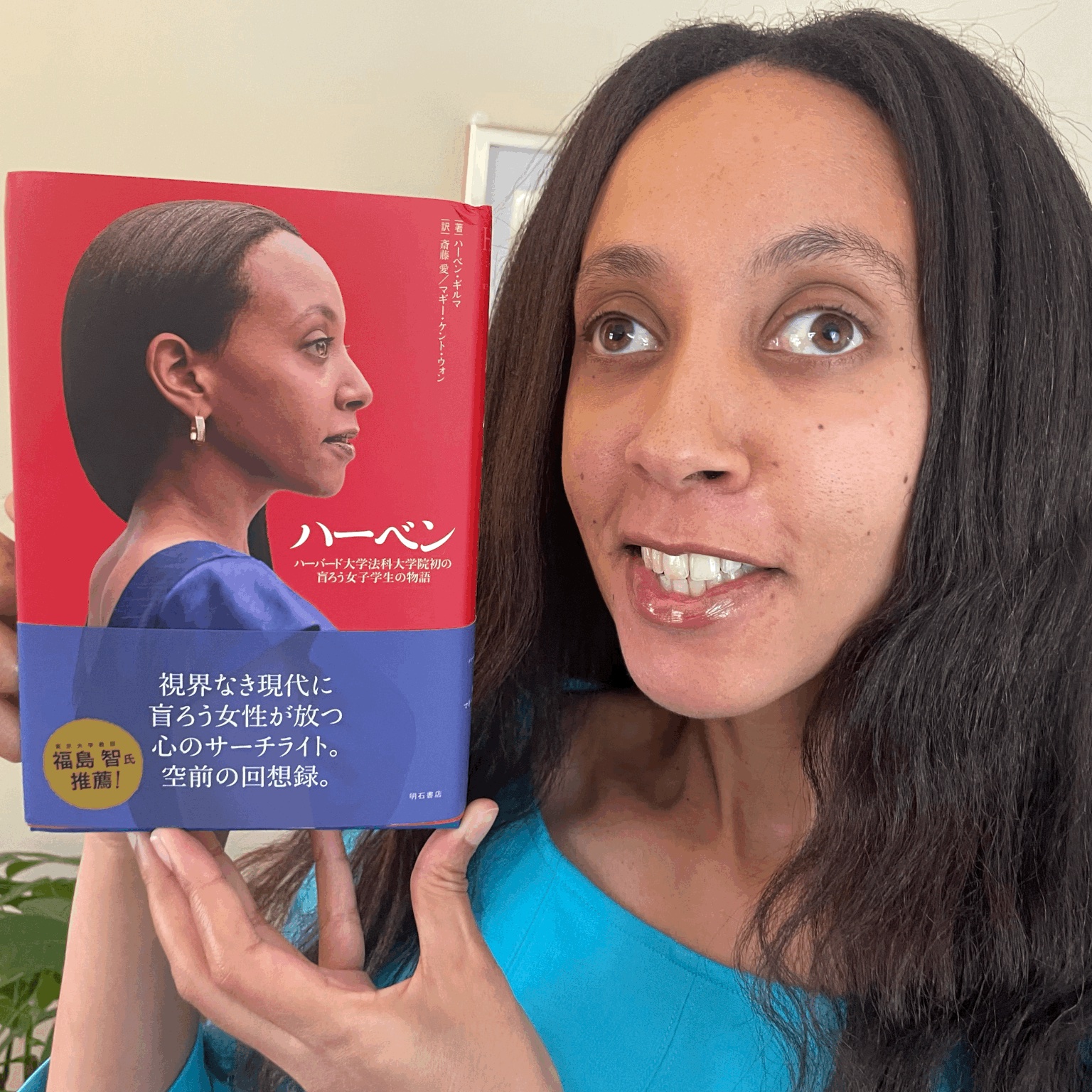 I’m smiling and holding up a book. The cover has a photo of me in profile, standing confidently in a blue dress. There is Japanese text along the side and bottom.