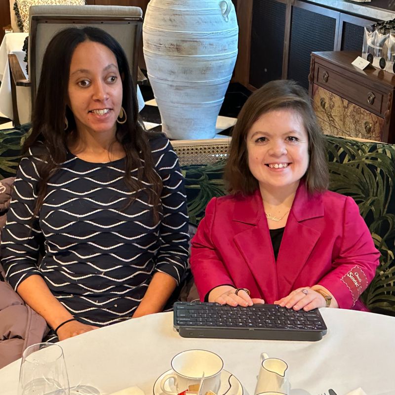Sinead, a visibly disabled white woman with shoulder-length brown hair wears a red coat with golden accents. Her hands are on a keyboard in front of her. Sitting beside Sinéad is Haben, a Black woman with long dark hair, gold earrings, and a blue and white dress. She’s reading braille on a computer that’s not visible under the table. They’re both smiling.