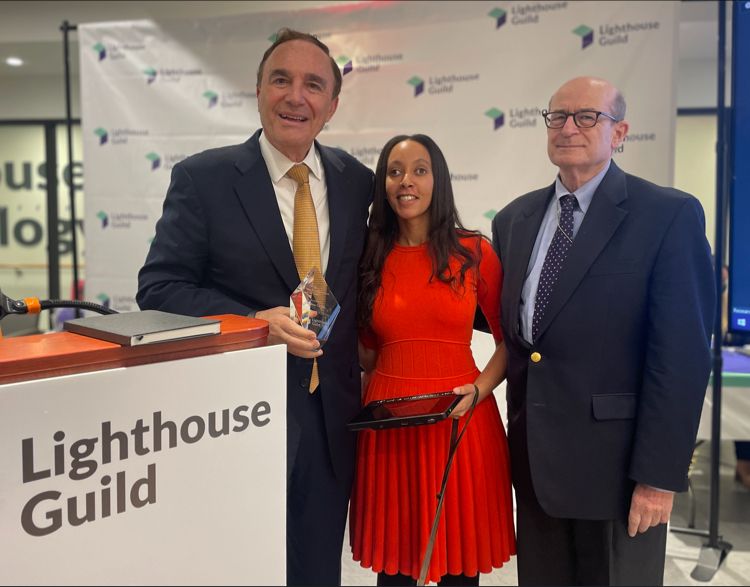 Three people standing by a podium that says Lighthouse Guild. Dr. Calvin W. Roberts, President & CEO of Lighthouse Guild, holds up a large glass, diamond-shaped award with my name on it. I’m standing in the middle, smiling and holding my braille computer. On my right stands Dr. Alan R. Morse, a professor at Columbia University’s Department of Ophthalmology and president emeritus of Lighthouse Guild.