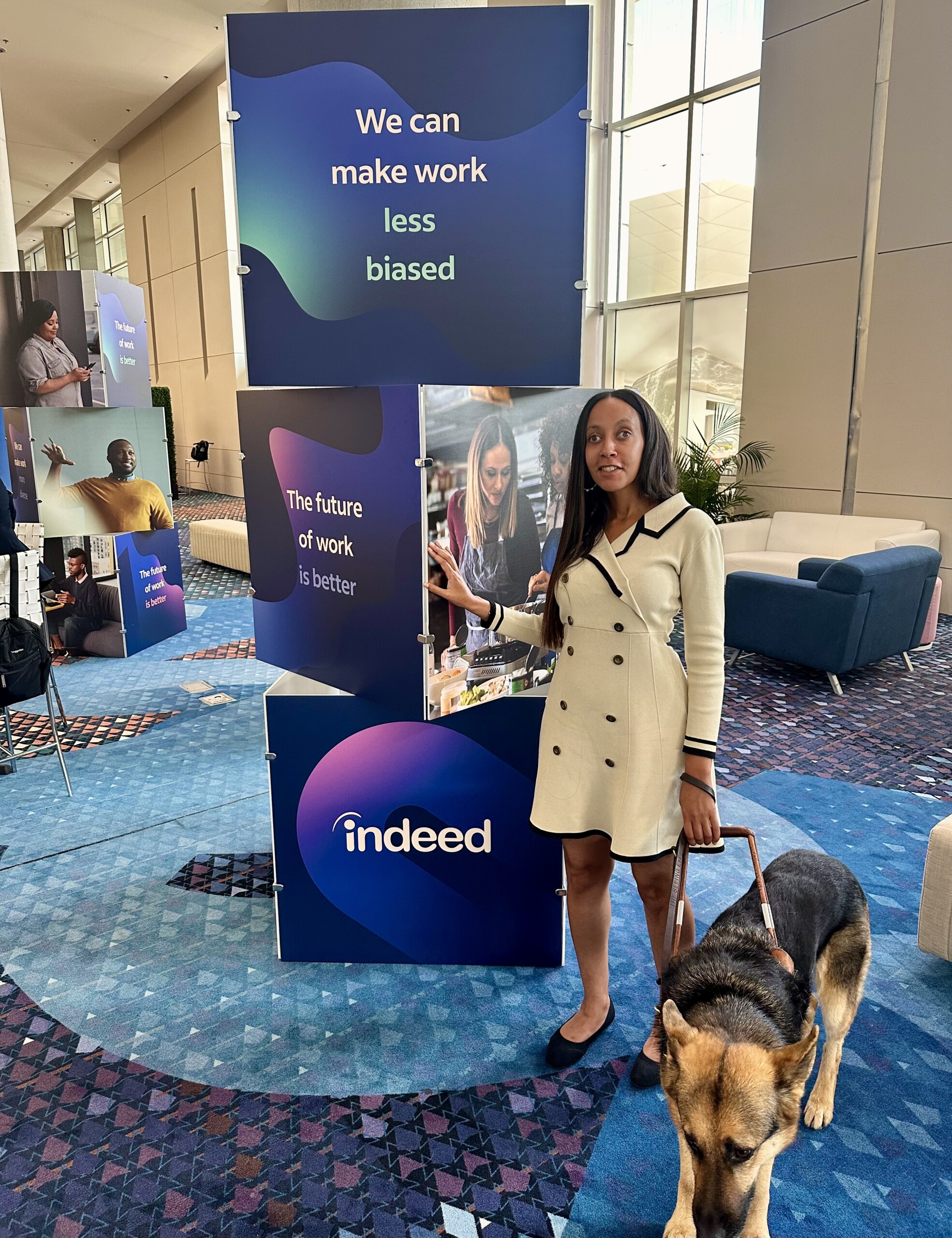 My Seeing Eye dog and I stand beside a tower of three, slightly rotated blocks. The bottom one has a purple background and says, “Indeed” in big white letters. The middle cube says, “the future of work is better” on one side and the other side has an image of three people cooking. The top cube says, “we can make work less biased.”