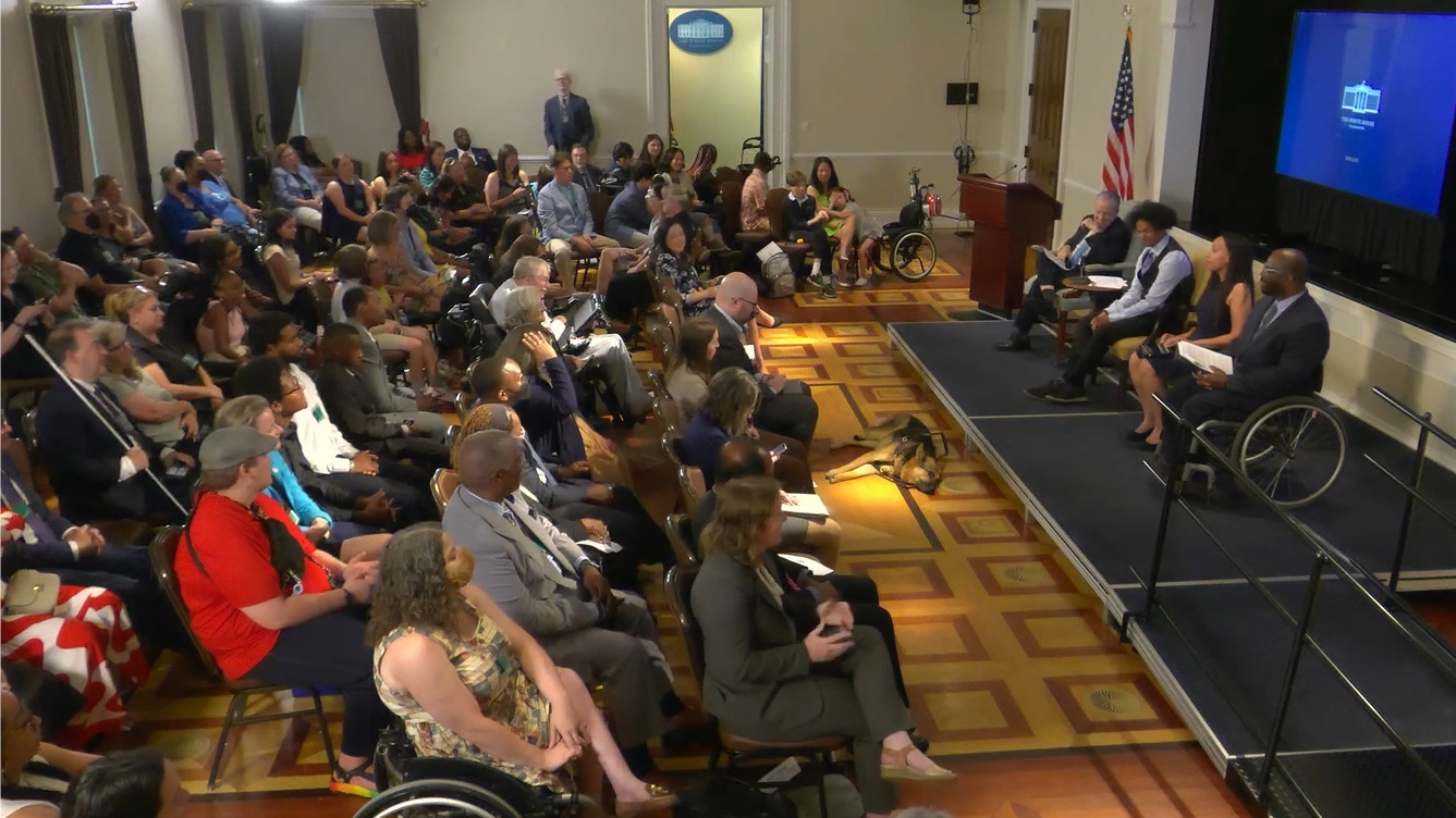 A crowded event space in the Eisenhower Executive Building. The audience is watching Haben Girma, Mychal Threets, Eric Harris, Gene Sperling, and Haben's guide dog Mylo onstage.