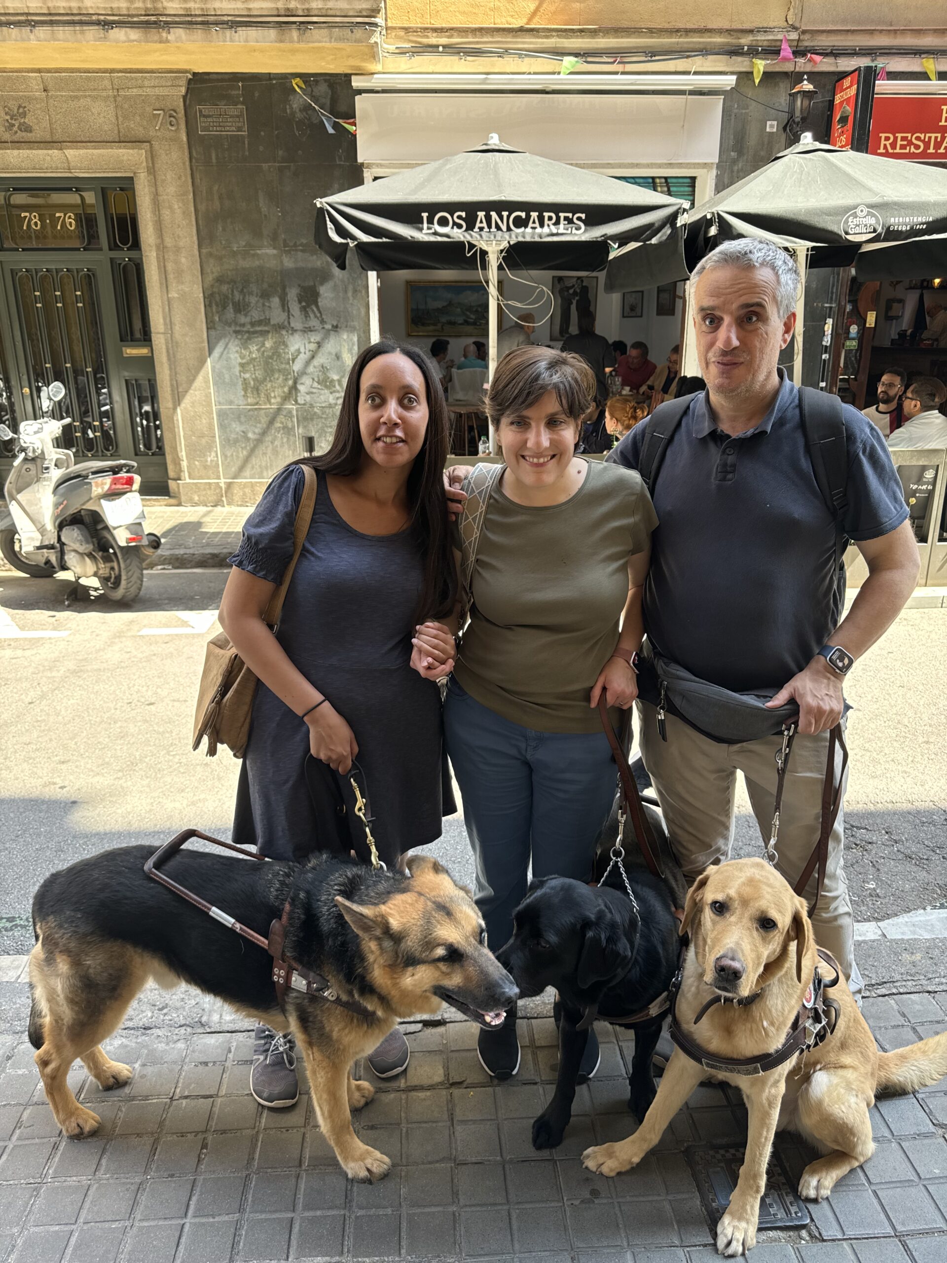 Haben, Daniela, and her husband José stand together on a sunny sidewalk. Haben wears a blue dress, Daniela wears a green shirt and jeans, and José wears a Navy blue shirt, khakis, and an Apple Watch. Next to German Shepherd Mylo is Rosie, a black lab, and beside José sits a yellow lab sweetly looking at the camera. Behind the smiling group, across the street, is a warm restaurant with indoor and outdoor seating.