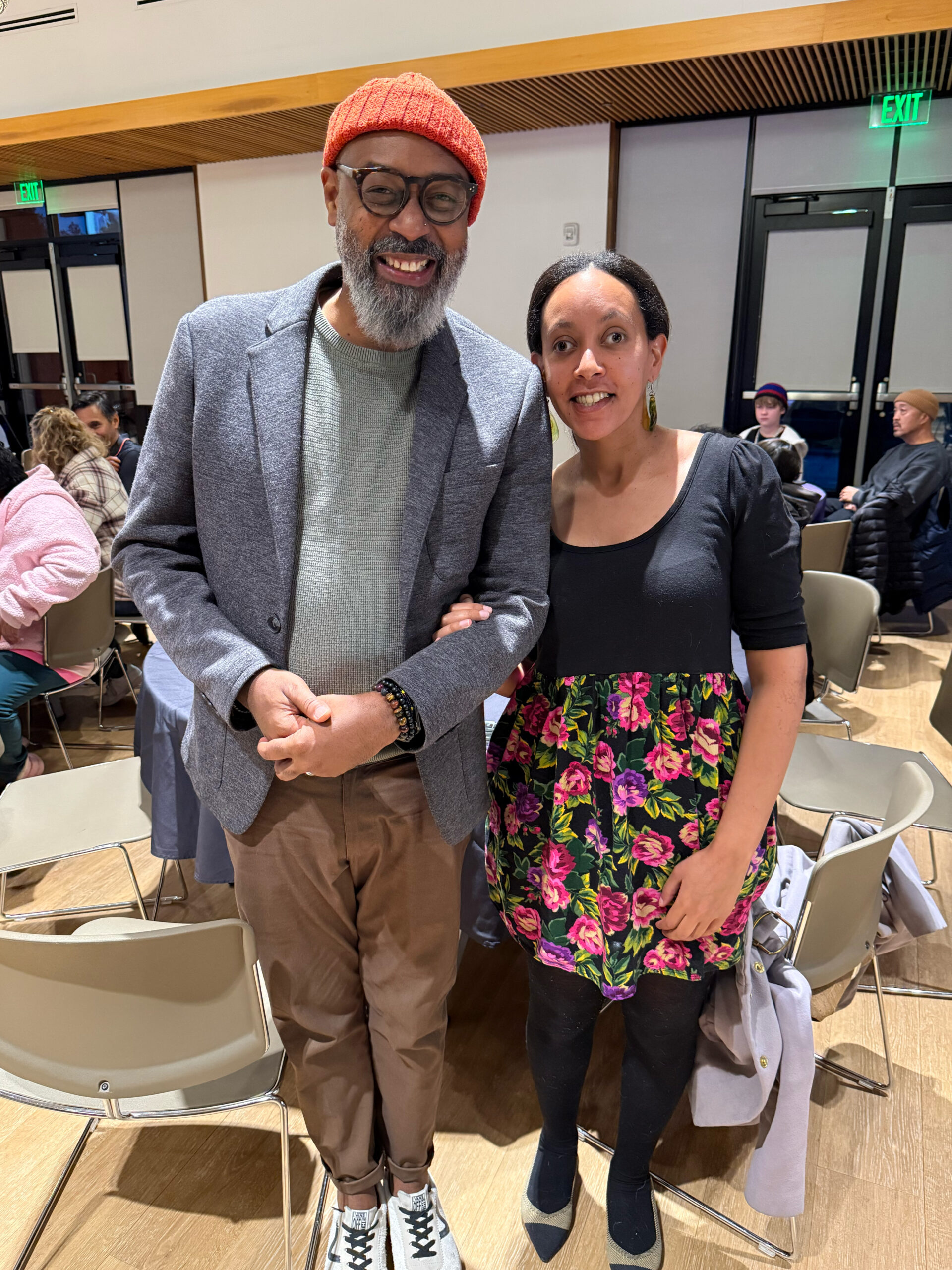 Haben stands beside Eric, a Black man in his fifties with glasses, a gray beard, and a warm smile. In the background other people are enjoying conversations around a large room full of tables and chairs.