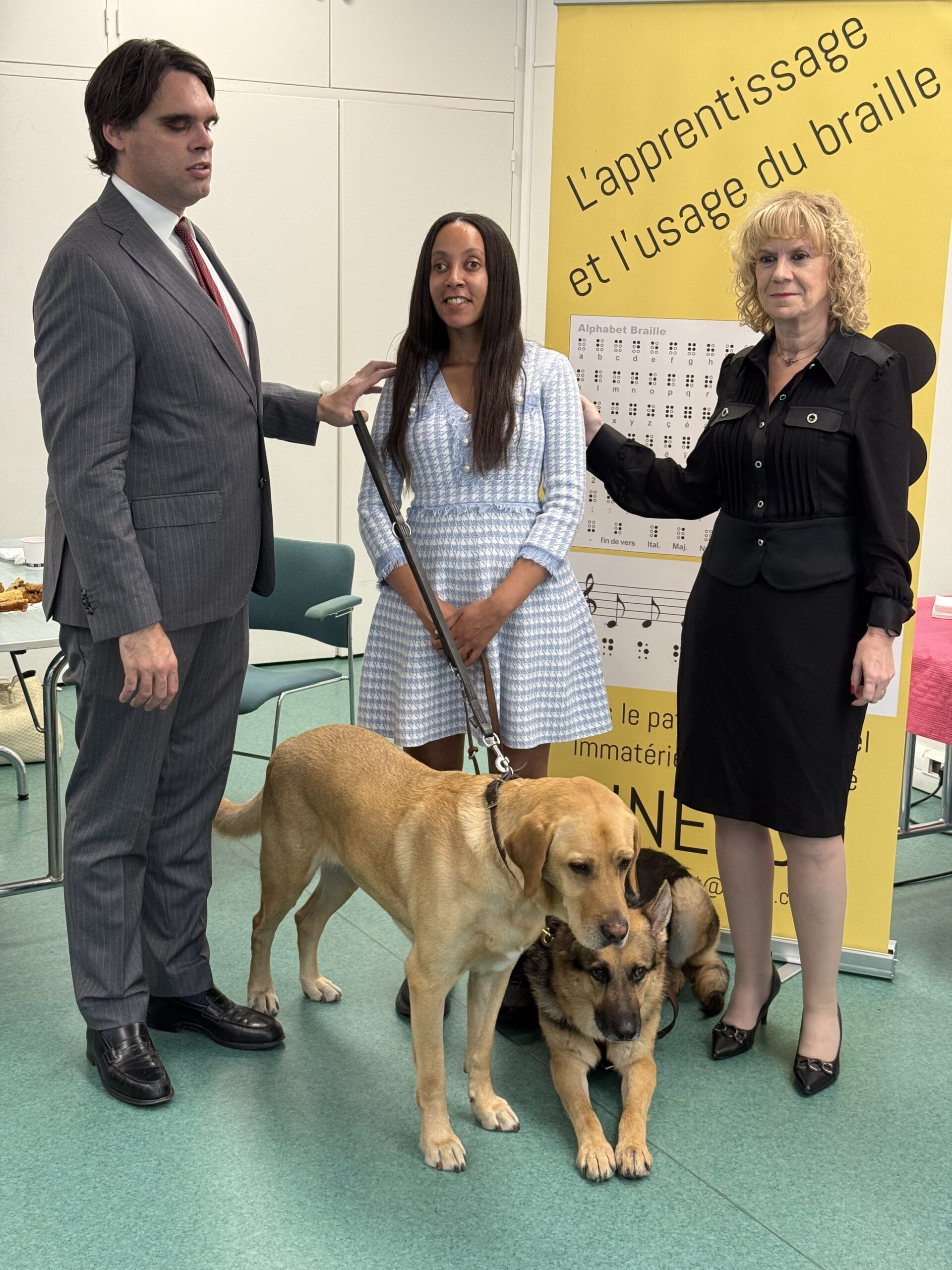President Matthieu Juglar and his guide dog Rider, Haben Girma and her guide dog Mylo, and Director General Carole Godin. They are standing in front of a vertical yellow banner with black text, “L’apprentissage et l’usage du braille.” A chart shows the Braille and print alphabets and numbers. Beside that, six large circles visually represent a standard Braille cell. The banner also shows Braille music notation.